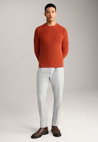 Maglione a coste rosso, pantaloni sartoriali grigi chiari e stivali in pelle marrone. Il maglione ha un collo a girocollo e un discreto logo sul manica.