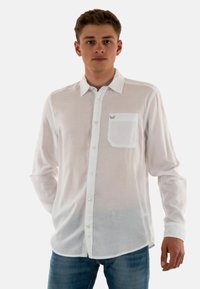 Chemise blanche à manches longues avec un devant à boutons, une seule poche poitrine et une texture uni, portée sur un jean bleu clair.