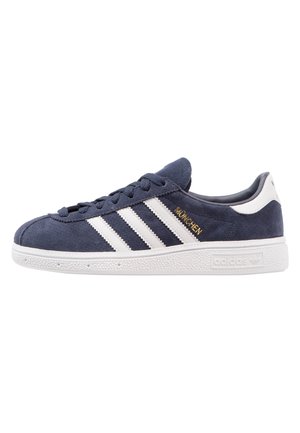 Navyblauwe suède sneaker met witte rubberen zool, witte Adidas-strepen, gouden tekst "MÜNCHEN" en een wit hielstuk.