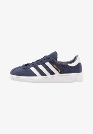 Navy blue suede sneaker with white rubber sole, white Adidas stripes, gold "MÜNCHEN" text, and white heel patch.