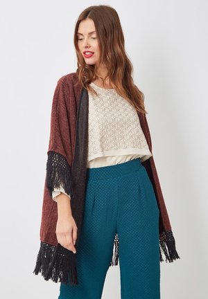 Mujer con cabello largo y castaño que lleva una blusa beige con estampado, pantalones texturizados en color verde azulado y un chal burdeos con flecos negros en los bordes.