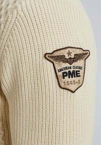 Crème gebreide trui met een textuurpatroon en een bruine geborduurde patch met vleugels, "American Classic PME," en het nummer 1549-A.
