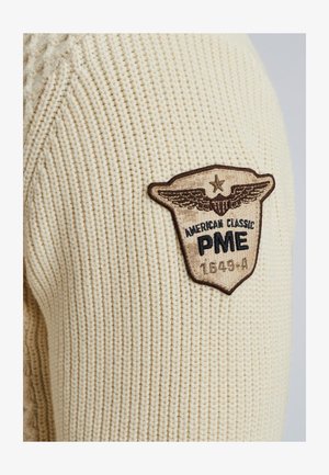 Maglione a maglia crema con un motivo testurizzato e una toppa ricamata marrone con ali, "American Classic PME," e il numero 1549-A.