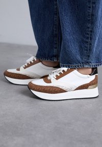 Hvit og brun sneaker med teksturerte detaljer, som har en tykk hvit såle, brune skinn-detaljer og en gullmetallisk hæl.