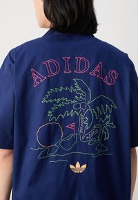 Person som bär en marinblå skjorta med broderad rosa "ADIDAS" text, gröna palmträd, golfflagga och guld Adidas-logga på ryggen.