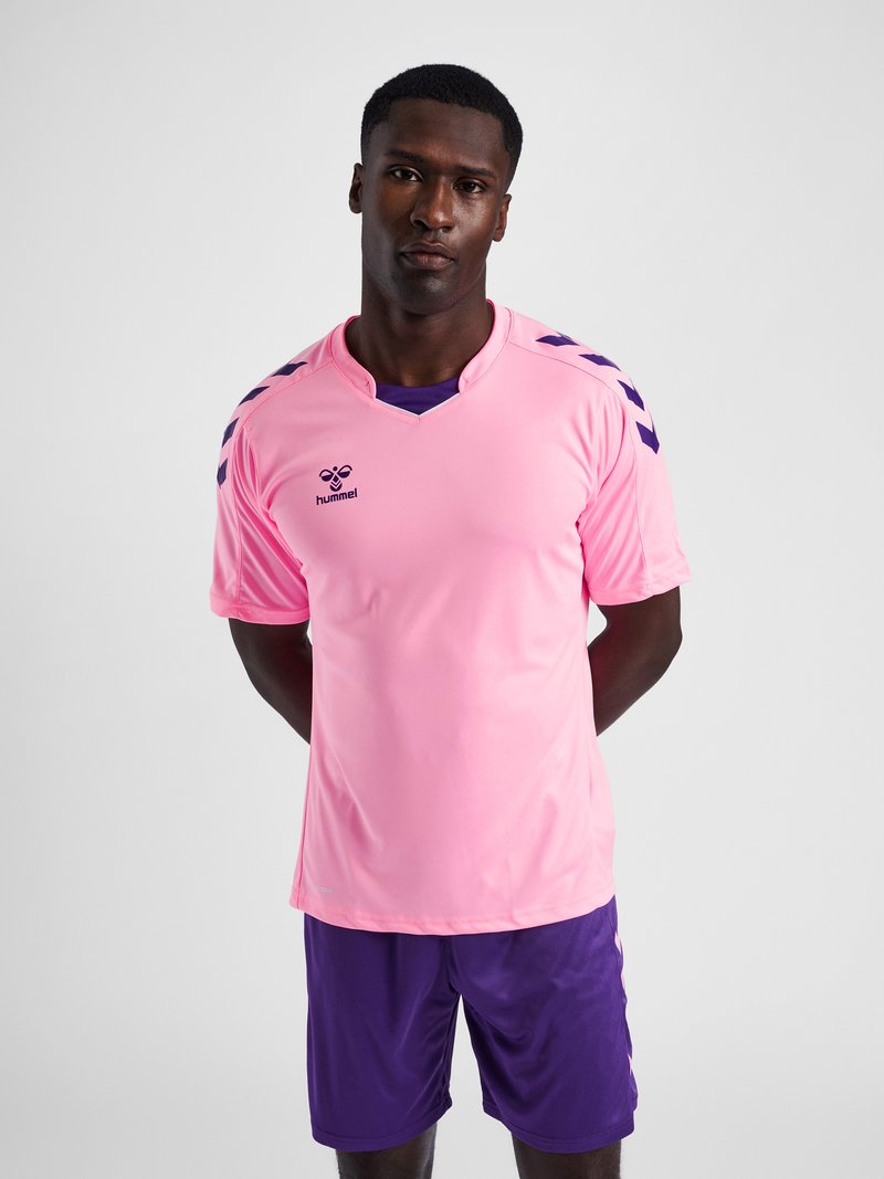 Homme portant un maillot de sport Hummel rose avec des bandes chevrons violettes sur les épaules et un short violet assorti, debout les mains derrière le dos.