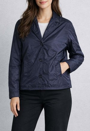 Donna con giacca blu navy abbottonata con revers sopra una camicia bianca, con una mano in tasca e pantaloni neri.