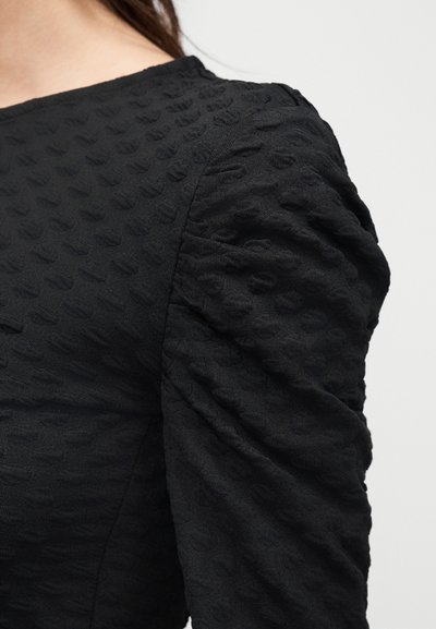 Κοντινό πλάνο στον ώμο ενός ατόμου που φοράει μαύρο ρούχο με υφή και φουσκωτά μανίκια, με στρογγυλή λαιμόκοψη, μπροστά σε απλό φόντο.