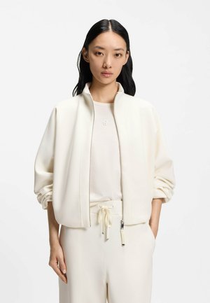 Cremefarbene Reißverschlussjacke aus weichem Stoff mit strukturierten Schultern, weiten Ärmeln und tiefen Taschen, kombiniert mit einem passenden Top und einer Hose.