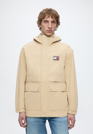 Uomo che indossa giacca beige con cappuccio, tasche frontali con patta e logo Tommy Jeans, abbinata a jeans blu, in piedi su sfondo bianco.