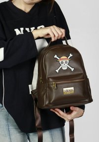 Mochila marrón de cuero sintético con forma redondeada, que presenta un diseño de calavera y huesos cruzados. Incluye un bolsillo con cremallera en la parte delantera y correas de tela para los hombros.