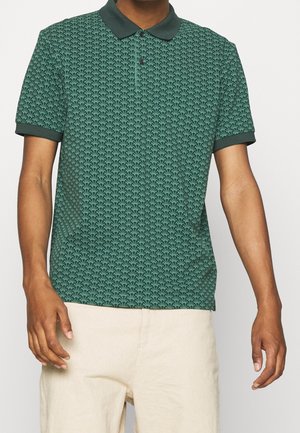 Homme portant un polo vert à motifs avec un col et des bordures noirs, associé à un pantalon beige, sur un fond blanc uni.