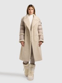 Beige dobbeltradet frakke med blødt, tekstureret stof og puffede ærmer; båret over en lys rullekrave og tætsiddende beige bukser, kombineret med lyse støvler.