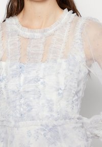 Robe blanche avec superposition en dentelle transparente, ornée d'un motif floral en bleu clair. Encolure et manches texturées avec des accents froncés.