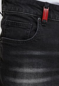 Vaqueros de mezclilla negra con un bolsillo frontal, una etiqueta roja cerca de la cinturilla y patrones de desgastado sutiles a lo largo de la tela.