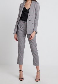 Kostým s kostkovaným vzorem, který obsahuje přiléhavý blazer a úzké kalhoty, vyrobený z měkké látky, s černými akcenty a lodičkami na kotníkovém pásku.