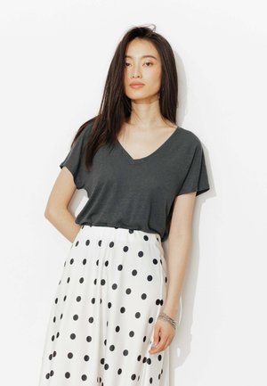 Femme aux longs cheveux foncés portant un T-shirt gris foncé à col en V et une jupe blanche à pois noirs, debout devant un fond blanc.