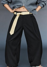Personne portant un pantalon noir ample avec une ceinture jaune clair nouée et un pull gris court zippé, mains sur les hanches, sur fond gris.