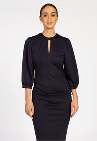 Frau, die ein eng anliegendes schwarzes Kleid mit dreiviertellangen Puffärmeln und einem vorderen Schlitz-Ausschnitt vor einem weißen Hintergrund trägt.