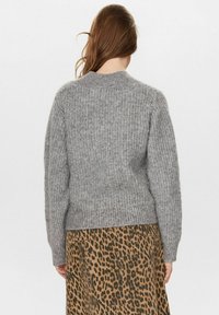 Žena s dlouhými hnědými vlasy, oblečená v texturovaném šedém pleteném svetru a hnědé sukni s leopardím potiskem, otočená zády k fotoaparátu.