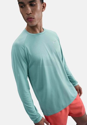 Junger Mann, der ein helltürkisfarbenes langärmliges Sportshirt und korallenrote Shorts trägt, blickt nach unten und leicht zur Seite.