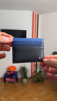 Μπλε και μαύρη θήκη καρτών με ραβδωτή υφή, που διαθέτει θήκες για κάρτες και λογότυπο. Κατασκευασμένη από δέρμα, κρατιέται ανάμεσα σε δύο χέρια.