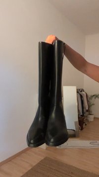Una mano tiene un paio di stivali alti in pelle nera in una camera da letto con uno specchio, un porta abiti e una pianta in vaso sullo sfondo.