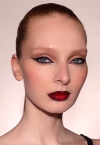 Nahaufnahme eines Gesichts mit glatter, heller Haut. Knallrote Lippen, definierter schwarzer Eyeliner und sanfte Augen-Make-up. Nach hinten gekämmtes Haar, neutraler Hintergrund.