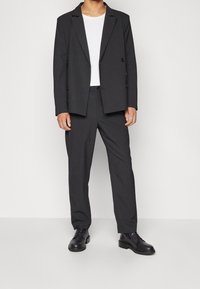 Mörkgrå skräddarsydd kostym med en rock med enkelknäppning och raka byxor, kombinerad med en vit t-shirt med rund hals och svarta skor.