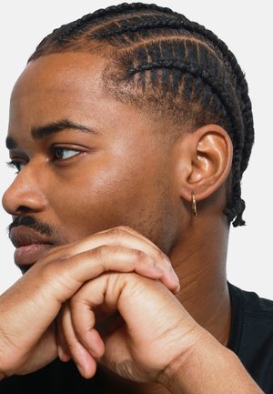 Braided hair with closely shaved sides, subtle highlight, gold hoop earring, smooth skin texture, hands clasped in front, dark shirt.