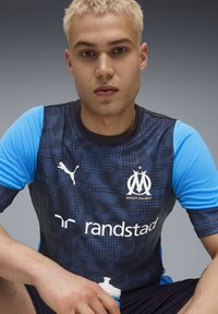 Maillot de sport Puma avec un design à motifs bleu foncé et des manches bleu clair. Logos affichés sur la poitrine, shorts noirs et une bouteille d'eau blanche.