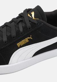 Crni suknjaški tenisice s bijelim kožnim detaljima i bijelom gumiranom potplatom. Sadrži zlatni logo i crne vezice. Kratki pogled sa strane.