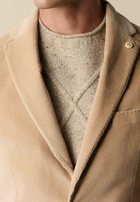 béžový kordový blejzr s širokými klopami, knoflíkovým detailem a náprsní kapsou, vrstvený přes texturovaný béžový pletený svetr s křížovým vzorem.