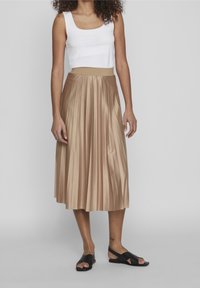 Woman wearing a white sleeveless top, a shiny beige pleated midi skirt, and black cross-strap sandals, standing against a plain background.