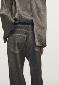 Pokročilá dlouhá halenka s vrásčitou strukturou zasunutá do džínů s vysokým pasem tmavé barvy, viditelnými zadními kapsami a detaily švů.