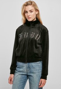 Chaqueta de felpa negra con cremallera y cuello alto, puños acanalados. Presenta "FUBU" en strass a lo largo del pecho. Combinada con jeans azul claro.