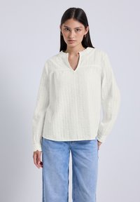 Blusa bianca a maniche lunghe con un design testurizzato, dotata di scollatura a V e dettagli arricciati, abbinata a jeans a gamba larga di colore azzurro chiaro.
