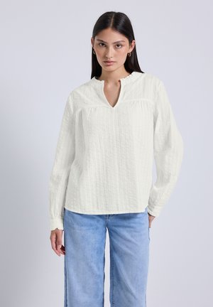 Weiße Bluse mit langen Ärmeln und texturiertem Design, ausgestattet mit einem V-Ausschnitt und gerafften Akzenten, kombiniert mit hellblauen, weiten Jeans.