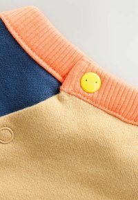 Kortærmet strikbluse med navyblå og ferskenfarvet ribbet krave samt en gul smiley-knap. Stoffet er blødt med en struktureret beige bund.