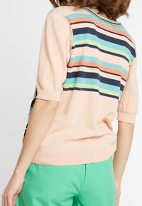 Gebreide pullover in zacht perzik met horizontale meerkleurige strepen, korte mouwen en een ronde hals. Groene broek zichtbaar.