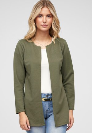 Giacca texture verde oliva, con scollo rotondo e apertura frontale. Maniche lunghe e vestibilità rilassata, abbinata a una maglietta bianca e jeans.