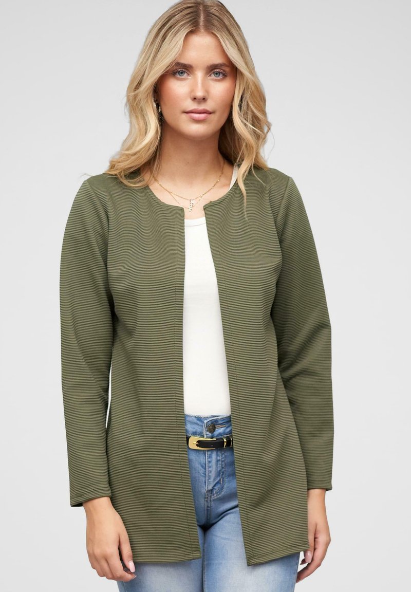 Olive green, textured jacket with a round neckline and open front. Long sleeves and a relaxed fit, paired with a white top and jeans.