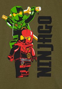 Grønne og røde ninja-figurer er avbildet på en oliven bakgrunn, med teksten "NINJAGO" i fet svart. Designet inkluderer sverd og karakteristiske kostymer.