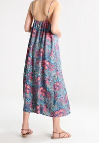 Robe maxi à fleurs bleue avec des fleurs roses et des feuilles vertes, présentant des bretelles fines et une coupe ample. Portée avec des sandales plates à lanières.