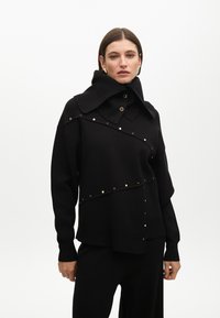 Suéter negro de punto con un cuello alto, detalles de broches dorados y un diseño asimétrico con un acabado texturizado. Estilizado con pantalones a juego.