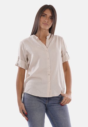 Giovane donna con capelli castani lisci che indossa una camicia beige con bottoni e maniche arrotolate e jeans blu, in piedi con una mano in tasca.