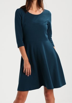 Femme portant une robe bleu sarcelle texturée, longueur genoux, manches trois-quarts et taille cintrée, debout avec une main dans la poche.