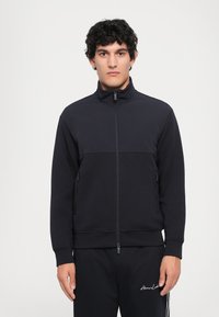 Svart zip-up treningsjakke i et mykt stoff med høy krage, med en horisontal søm, to sidelommer og glidelås.