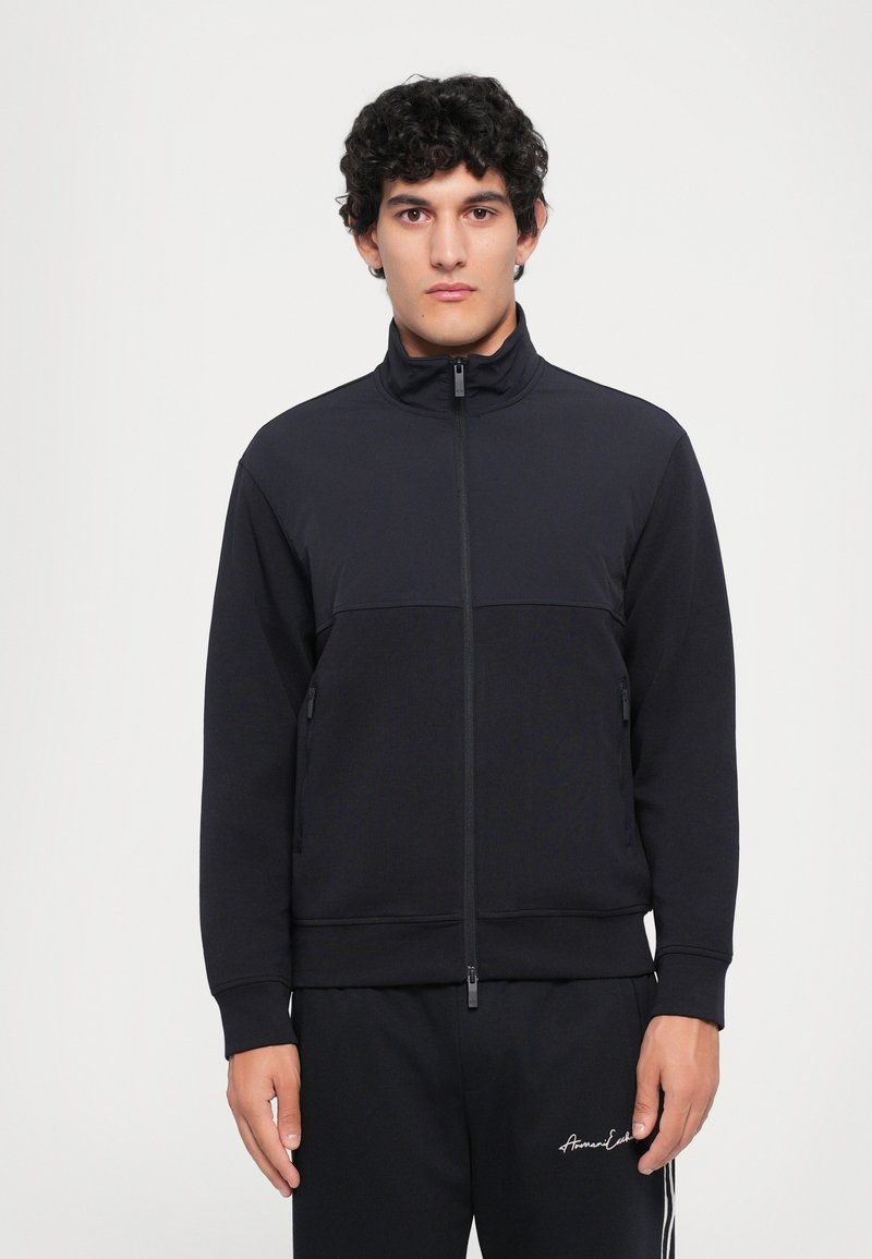 Svart zip-up treningsjakke i et mykt stoff med høy krage, med en horisontal søm, to sidelommer og glidelås.