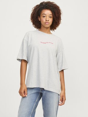 Giovane donna con capelli ricci, indossa una maglietta larga grigio chiaro con scritta rossa "Be Savage Not Average" e jeans blu sbiaditi, in piedi su sfondo bianco.